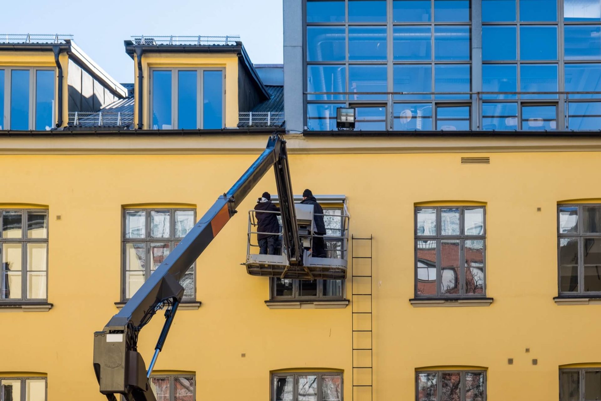 Arbetare ombord på en skylift tar sig an uppgiften att renovera fasaden på en gul byggnad, utrustad med stora fönster